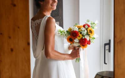Photographie de mariage : une mariée souriante en robe blanche élégante tient un bouquet de fleurs colorées avec des tournesols, des fleurs rouges et blanches. La scène est capturée près d’une fenêtre, avec son reflet visible dans un grand miroir en bois. Image réalisée par un photographe de mariage pour immortaliser un moment plein d’émotion et de naturel.